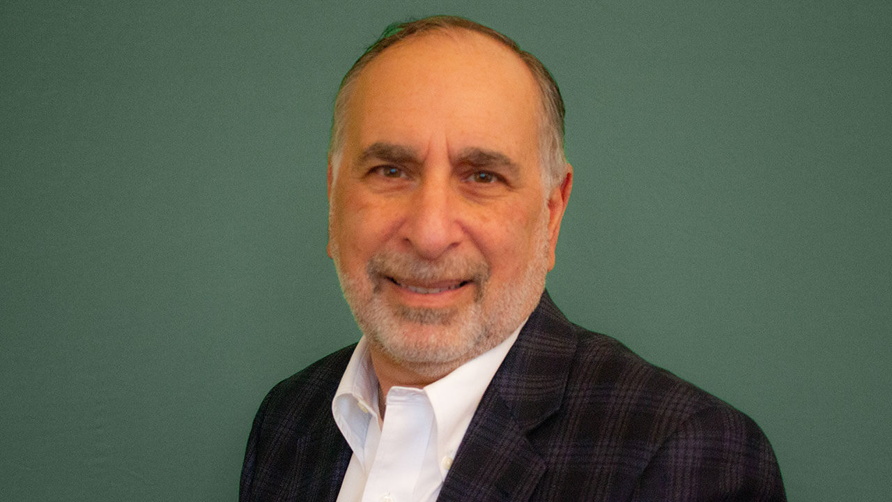 andy keller, a gray haired man with a gray beard smiles at the camera wearing a black blazer and a white button up in front of a green background